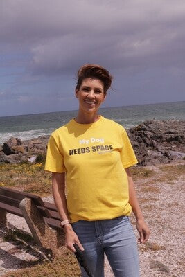 A smiling woman standing near a rocky ocean shoreline wearing a bright yellow t-shirt with the text "My Dog Needs Space". This t-shirt is ideal for dog owners whose pets may be anxious, unwell, elderly, nervous, adopted, or in training.