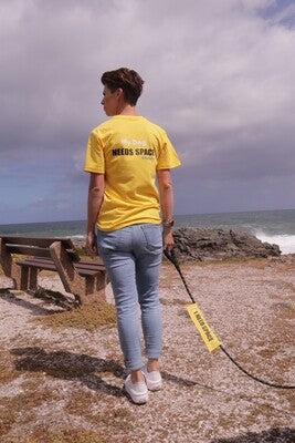 A woman standing near a rocky ocean shoreline wearing a bright yellow t-shirt with the text "My Dog Needs Space" with a matching yellow leash cover with the text "I Need Space" attached to a dog leash. This t-shirt is ideal for dog owners whose pets may be anxious, unwell, elderly, nervous, adopted, or in training.